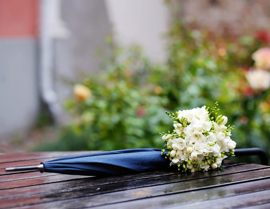 A closed blue umbrella and a bouquet of white flowers rest on a wet wooden bench, set against the blurred greenery. This serene scene reminds us that even amidst nature's beauty, avoiding wedding planning mistakes is key to ensuring your special day in Austin goes smoothly.