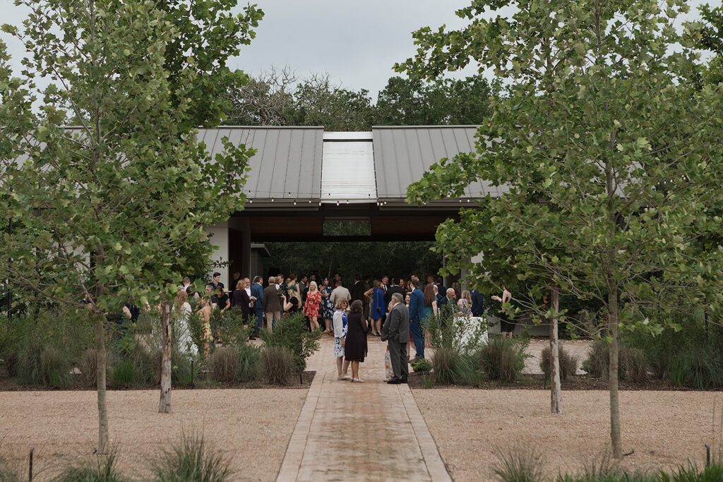 A group of people gather outside the Grand Lady, a modern building with a metal roof, elegantly surrounded by trees and plants.