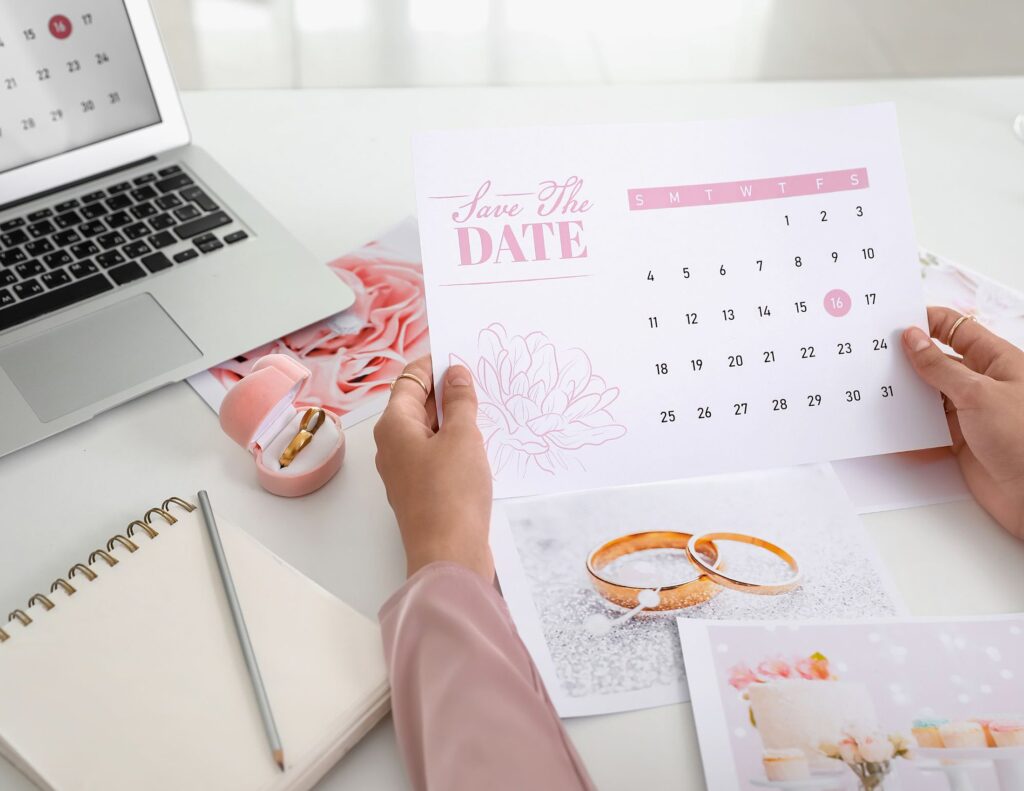 A person holds a "Save the Date" card with a calendar, surrounded by essentials like a laptop, notebook, stapler, and wedding ring images on the table—a scene reminding us of common wedding planning mistakes to avoid for your Austin celebration.