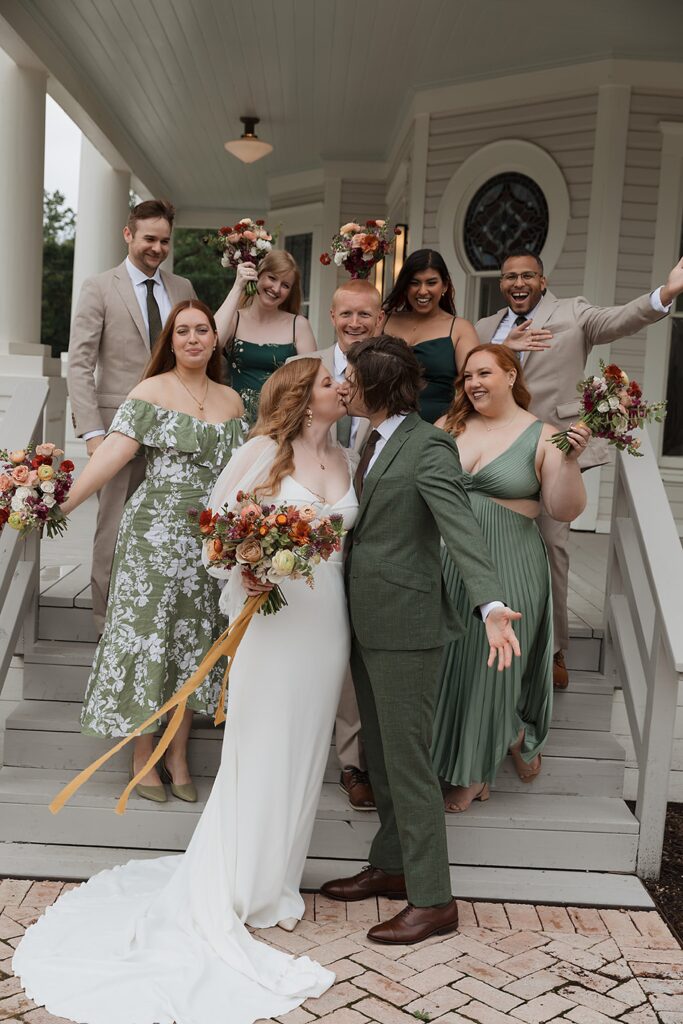 A couple kisses at a wedding on the steps of The Grand Lady. The bride wears a white dress and holds flowers, while the groom is in a green suit. Their bridal party, bridesmaids, and groomsmen surround them in joyous celebration.