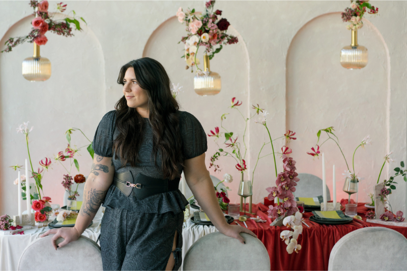 Sydney Meyer, Austin wedding planner, wears a dark dress and stands before a decorated table with red and pink flowers, candles, and tableware—an elegant setting that highlights the many reasons to hire a wedding planner for your special day.