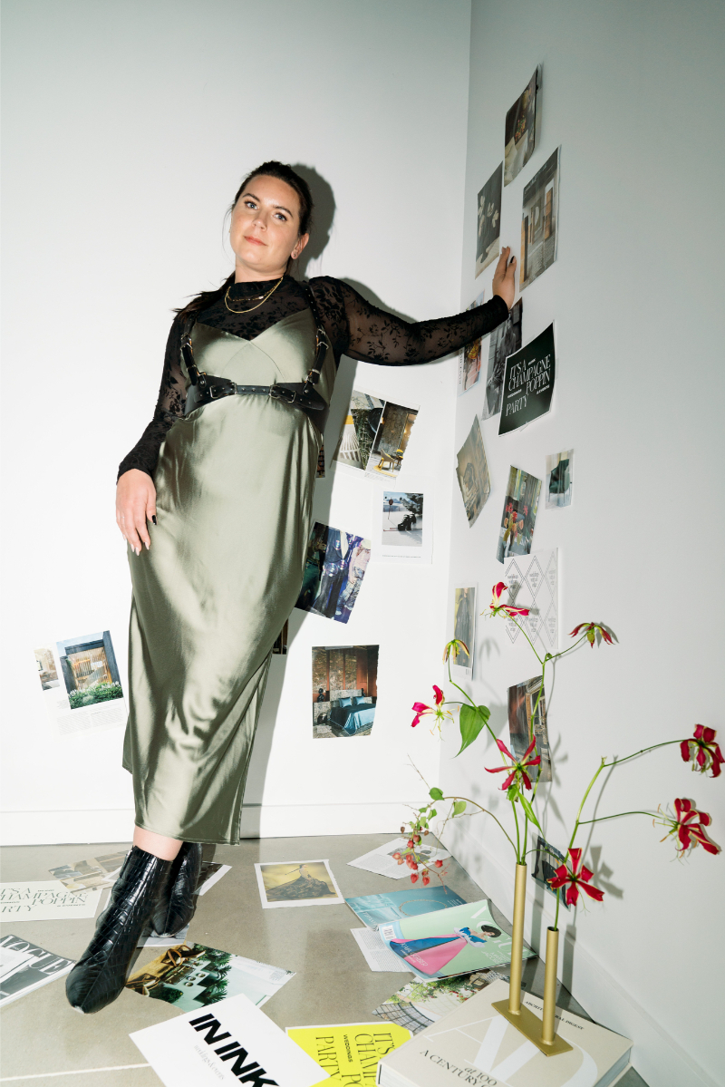 Sydney Meyer, Austin wedding planner of In Ink Weddings, wears a green satin dress and black boots stands in a corner decorated with art prints and posters; a vase with red flowers is nearby.