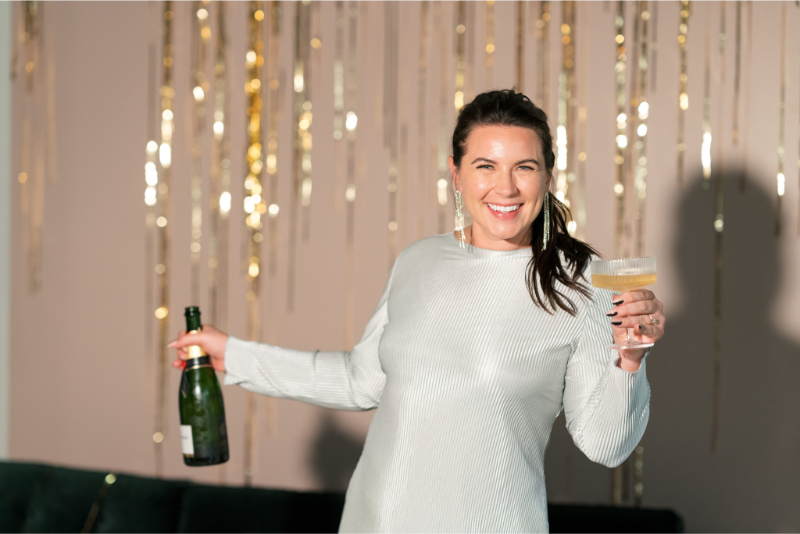 Sydney Meyer, Austin wedding planner, smiles, holding a glass and bottle of Dom Pérignon, with gold streamers in the background—capturing one of the many joyful moments and reasons to hire a wedding planner for your special day.