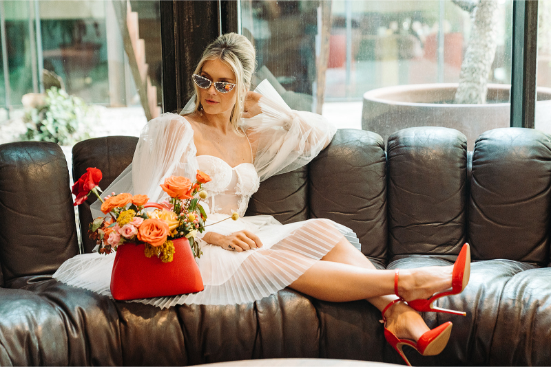 A stylish bride lounging on a dark leather sofa, wearing a white corset-style dress with dramatic tulle sleeves and vibrant red heels. She accessorizes with retro sunglasses and a bold red bag filled with bright orange, yellow, and pink flowers, capturing a chic and modern wedding look.