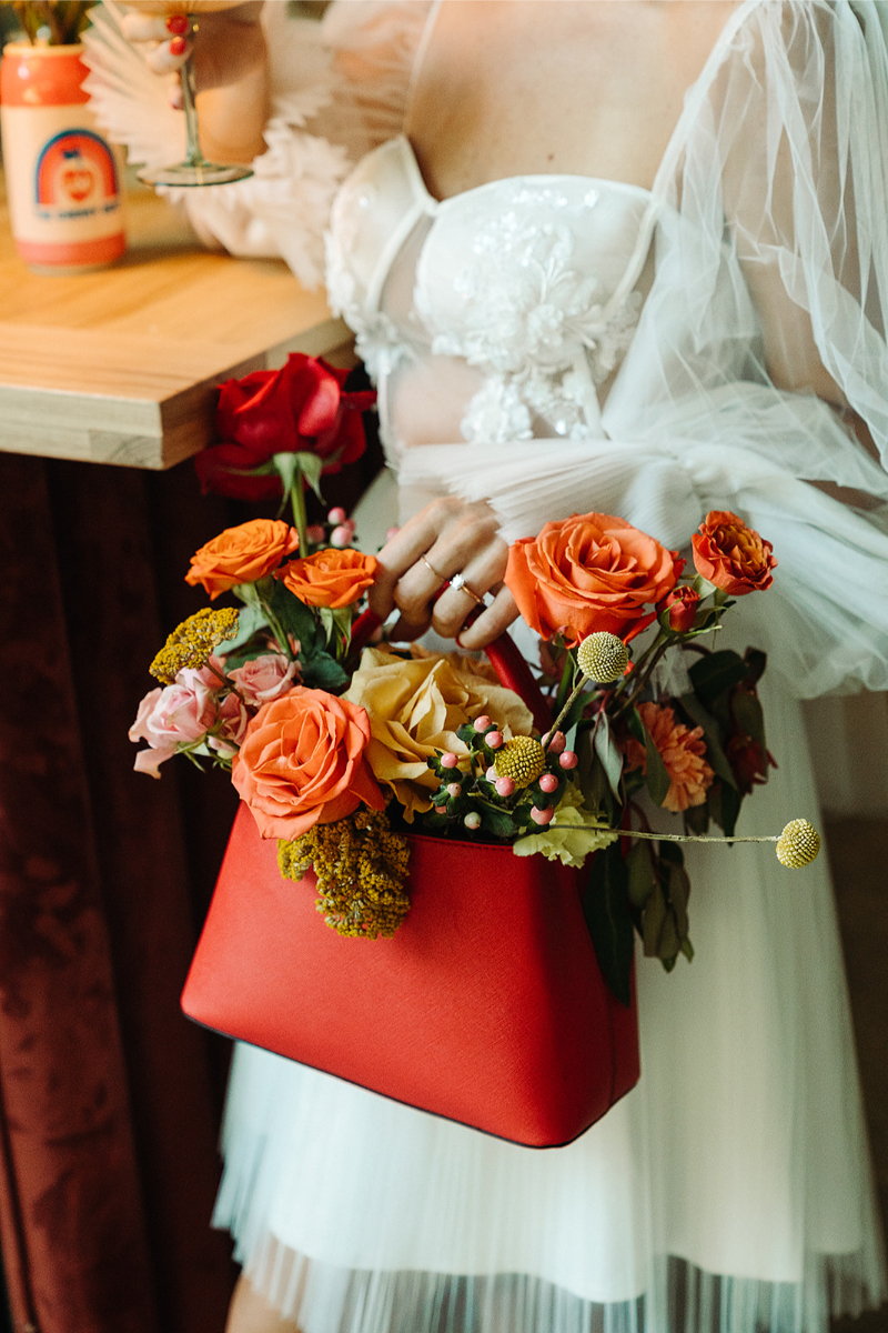 Close-up of a red handbag filled with vibrant wedding florals, including orange, yellow, and pink blooms, held by a bride wearing a white dress with intricate embroidery. This image highlights one of the standout 2025 wedding trends: bold and colorful details.