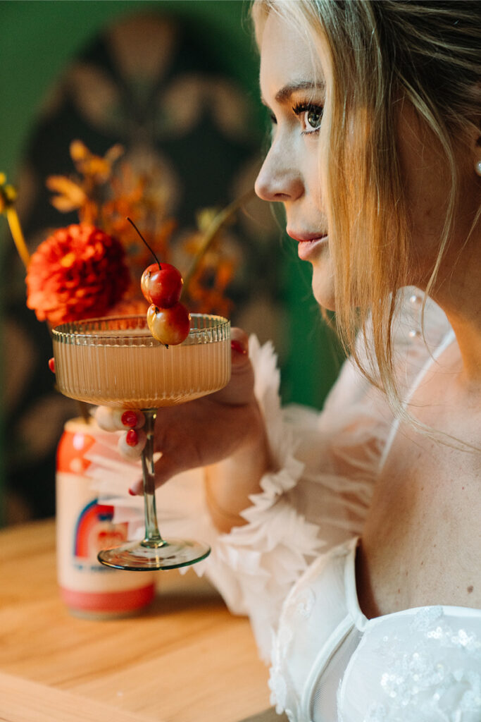 A close-up of a bride sipping a cocktail garnished with small cherries, set against a backdrop of a colorful bar.