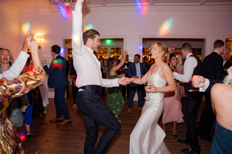 A joyful bride and groom dance enthusiastically on the reception floor, surrounded by vibrant party lights and celebrating guests.