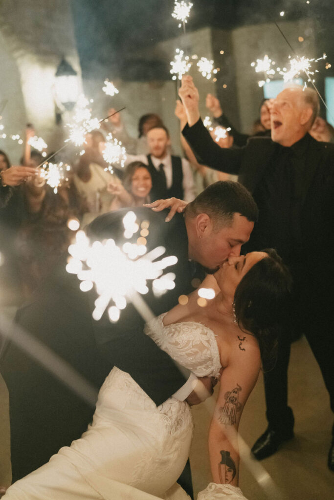 Newlyweds share a romantic kiss during a sparkler send-off, surrounded by joyful guests holding glowing sparklers.