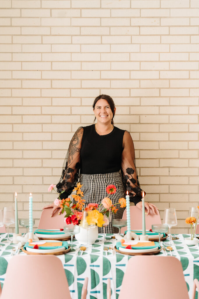 Sydney, owner of In Ink Weddings, standing behind a beautifully decorated table with vibrant floral arrangements, colorful tableware, and pastel pink chairs, as she discusses 2025 wedding trends. 