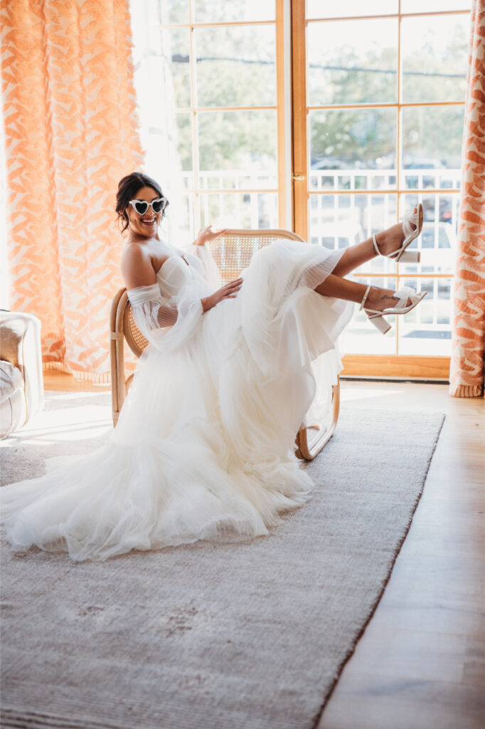 Bride in a white dress and sunglasses sits on a chair, with legs up, in a sunlit room with orange curtains.