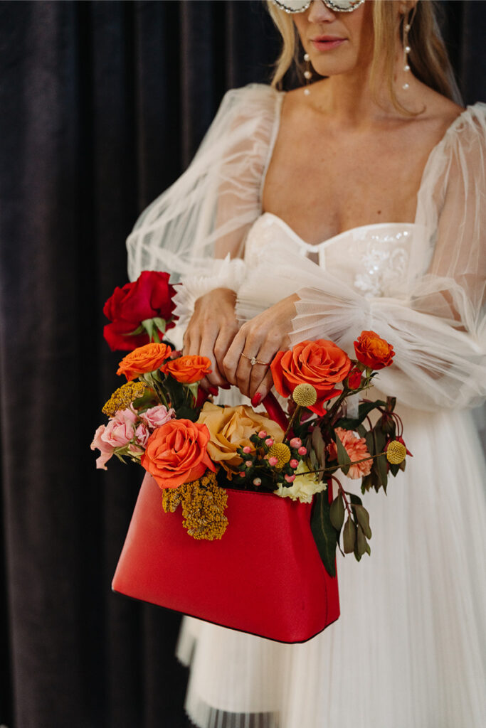 A bride in a white dress holds a red handbag filled with vibrant orange, pink, and yellow flowers, standing against a dark curtain backdrop.