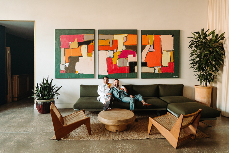 Two grooms sit on a dark green sectional at South Congress Hotel. There are abstract paintings on the wall above them, perfect for their art-inspired wedding.