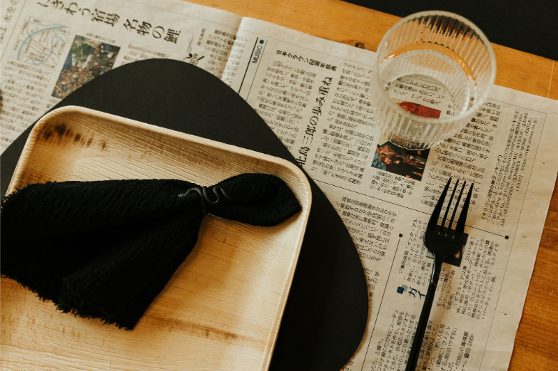 A wooden plate with a black cloth napkin, a glass of water, and a black fork on a newspaper place setting.