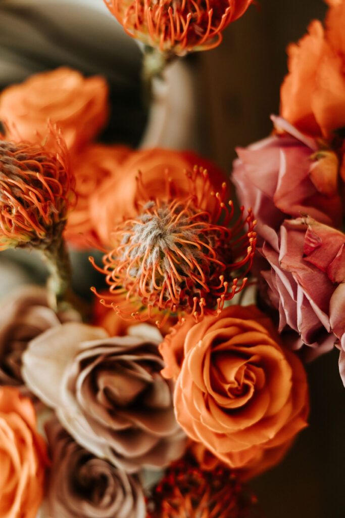 A close-up of a floral arrangement with orange roses, purple roses, and orange pincushion proteas.