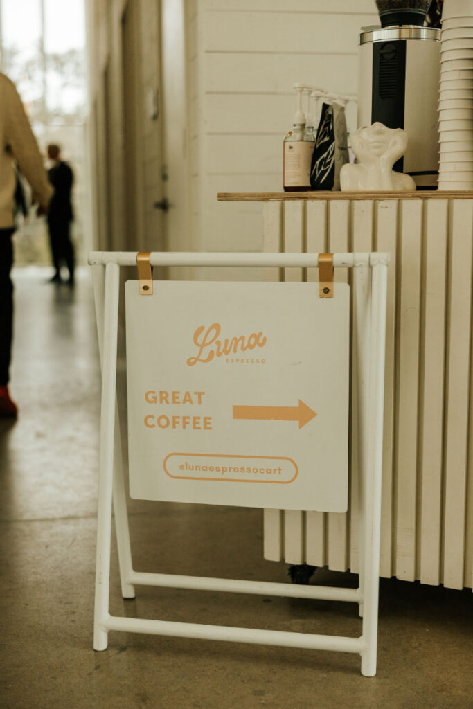 A sign reads "Luna Espresso" with an arrow and "Great Coffee." It is displayed on a stand near a counter with coffee cups and a dispenser.