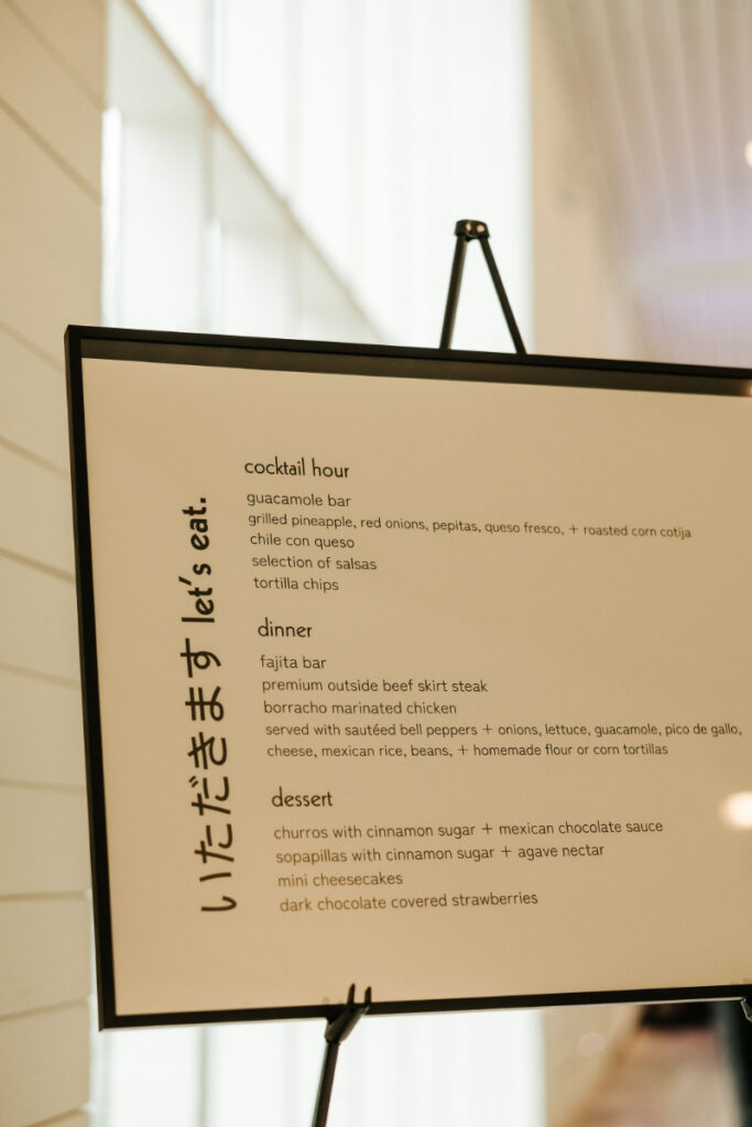 A menu on an easel with sections for cocktail hour, dinner, and dessert. Vertical text on the left reads "let's eat.