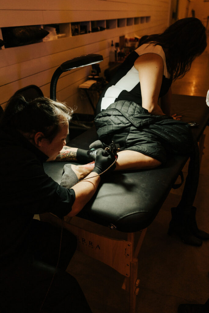 Person receiving a tattoo on their leg from a tattoo artist in a dimly lit room.