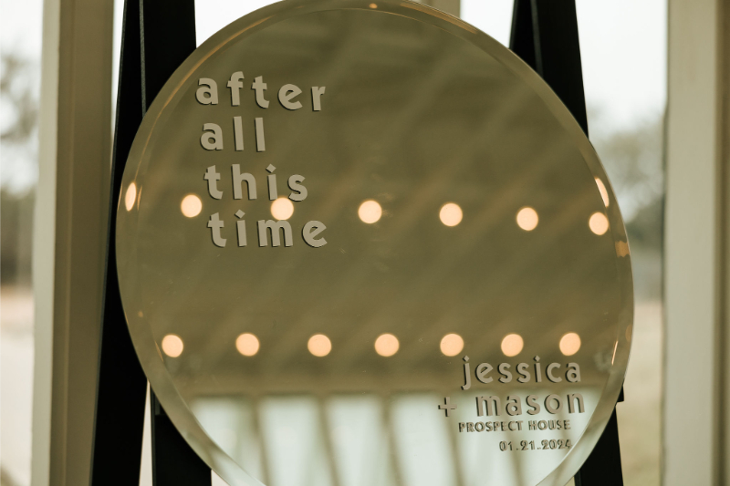 Round mirror with the text "after all this time" on the left and "jessica + mason prospect house 01.21.2022" on the bottom right, reflecting blurred lights.