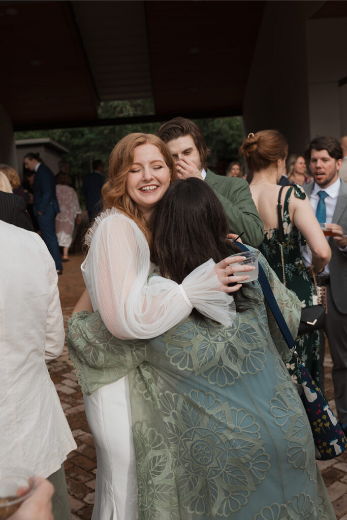 Two people embracing at an outdoor event, surrounded by others in formal attire. The person on the left is wearing a white dress, and the person on the right has a green patterned shawl.