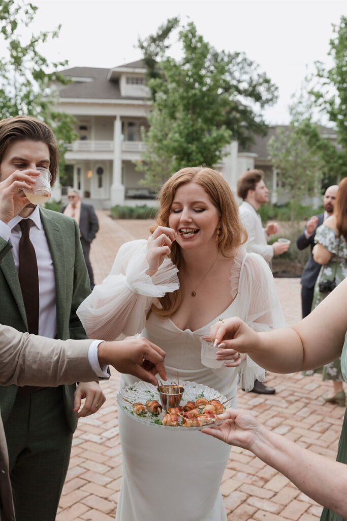 A bride in a white dress eats shrimp cocktail at an outdoor gathering. Other guests enjoy drinks and food around her. A large house is visible in the background.