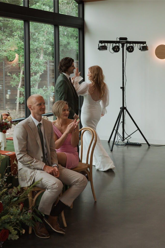 A bride and groom share a high-five at their wedding while two seated guests, a man and a woman, clap. There's a light stand in the background and a window showing greenery outside.