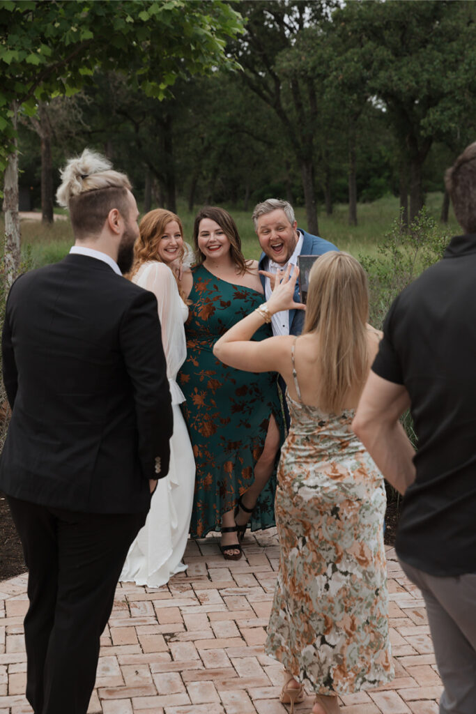 Group of people posing for a photo outdoors; one woman is taking their picture. There are trees in the background, and everyone is dressed formally.