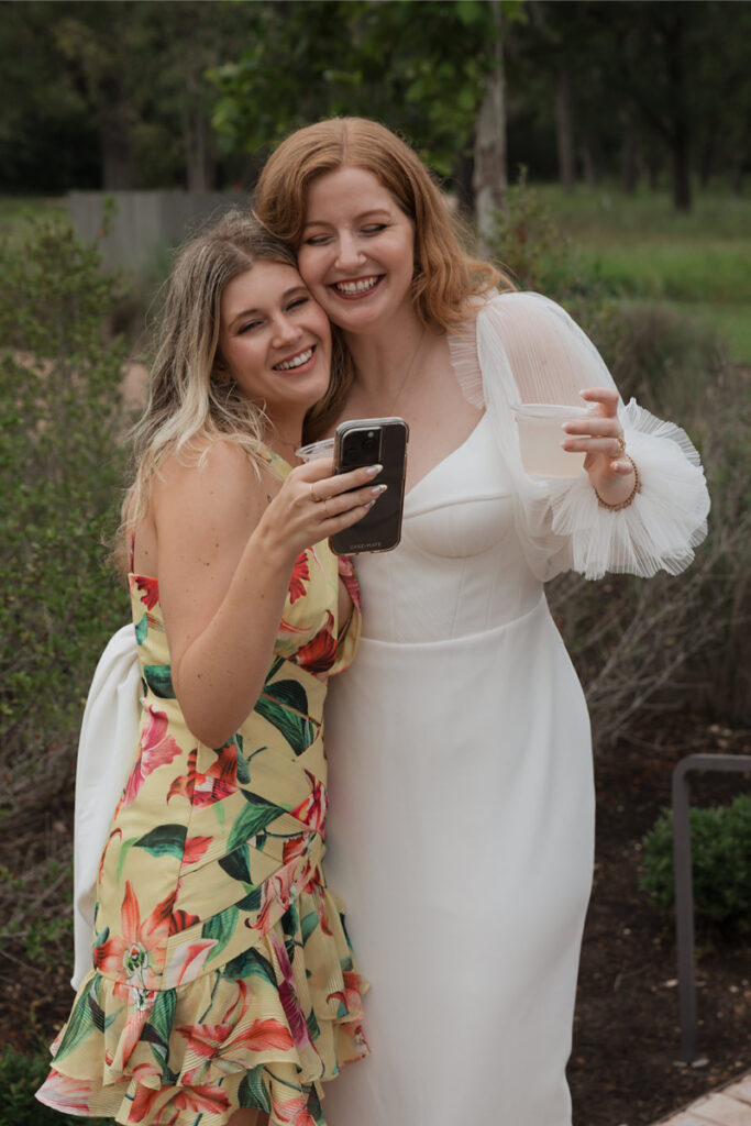 Two women smiling and looking at a phone. One is in a floral dress, the other in a white dress holding a cup. They stand outdoors with greenery in the background.