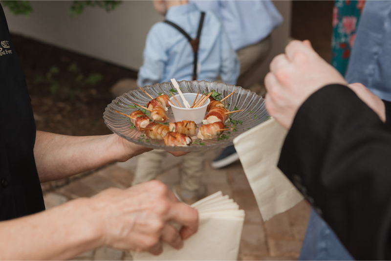 Person holding a plate with bacon-wrapped appetizers and a dipping sauce. Another person holds napkins. People in casual attire are in the background.