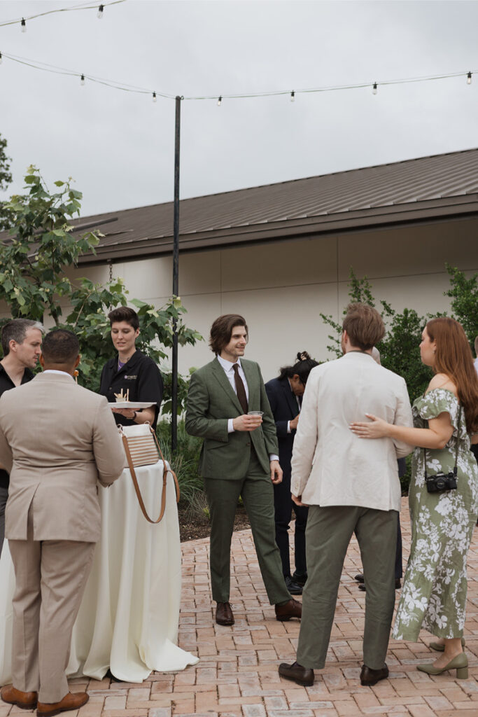 A group of people in formal attire standing and talking outdoors near a round cocktail table with a white tablecloth. One man in a green suit holds a drink.