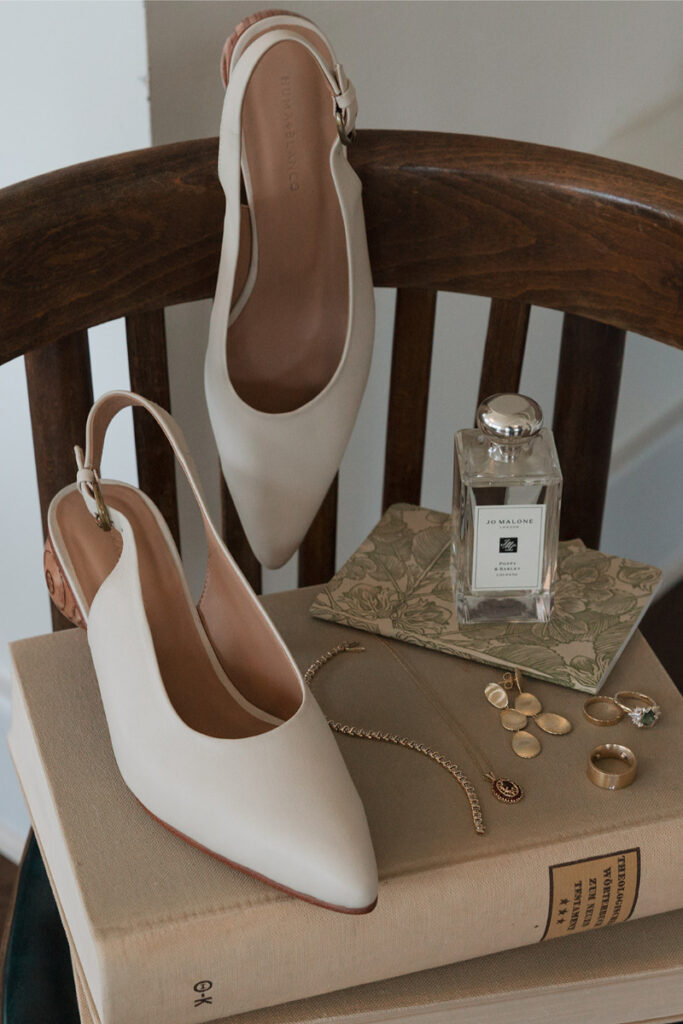 Beige pointed-toe shoes on books, with a perfume bottle, jewelry, and floral-patterned notes on a wooden chair.