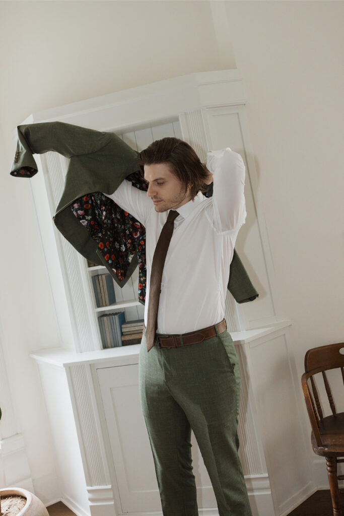 A man in a white shirt and green pants puts on a green jacket with a floral lining, standing in a room with a bookcase and a wooden chair.