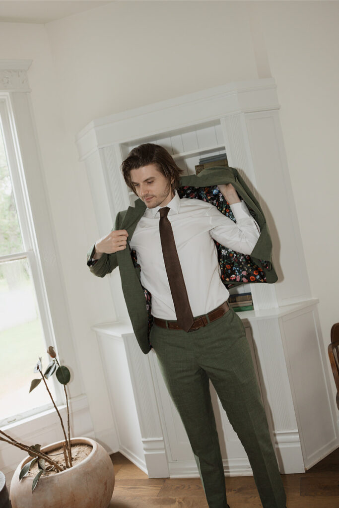 Man in a white shirt and tie adjusts his green suit jacket near a window. A potted plant on a wooden floor is visible.