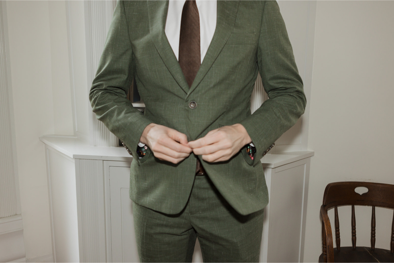 Person in a green suit adjusts their jacket in a room with a wooden chair and white walls.