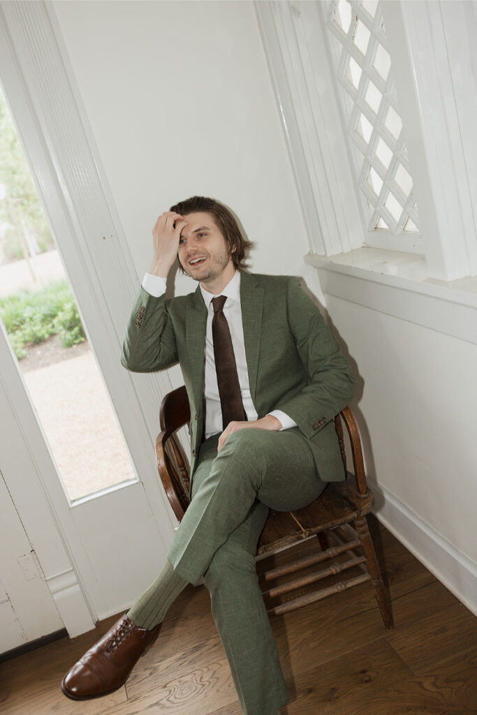 Man in a green suit and brown shoes sits on a wooden chair by a window, touching his forehead and smiling.