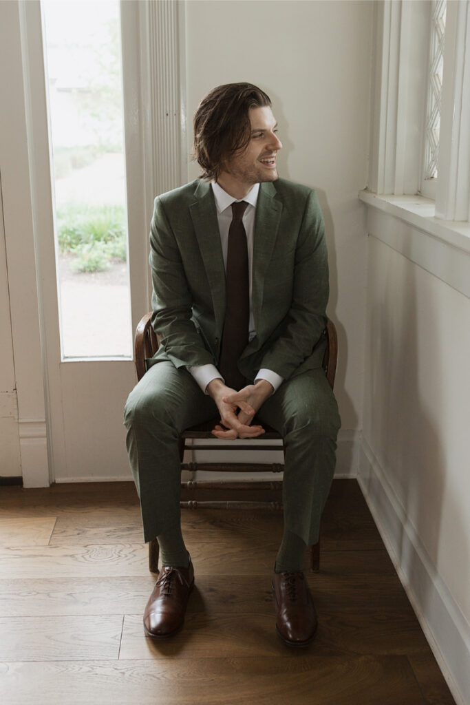 A man in a green suit and brown shoes sits on a chair by a window, looking to his left, smiling.