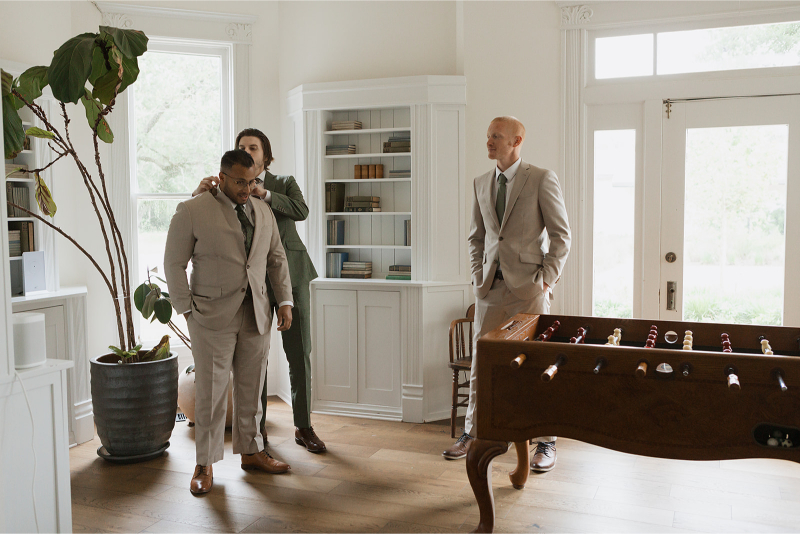 Three men in suits stand in a bright room with bookshelves and a foosball table. One adjusts another's tie while the third looks on. A large potted plant is nearby.