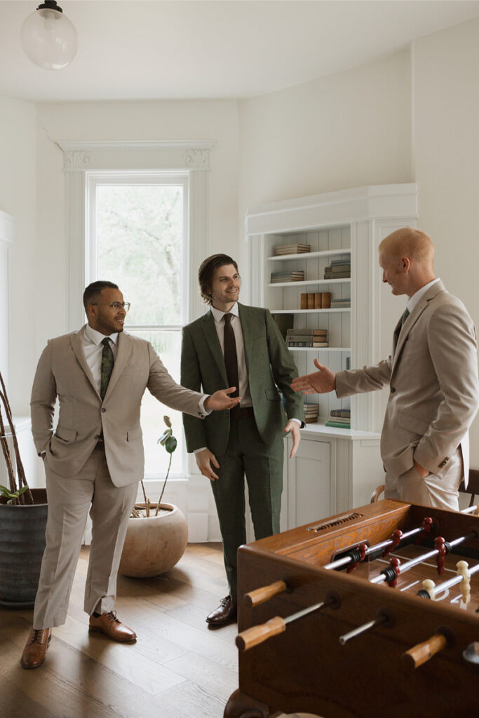 Three men in suits are standing in a room with a foosball table, conversing near a window and bookshelves.