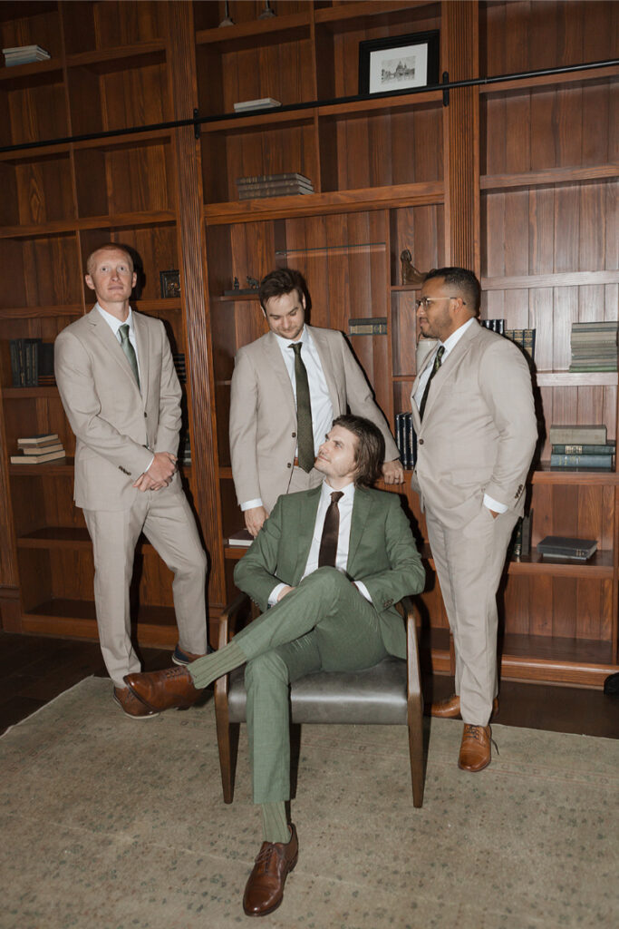 Four men in suits pose in a wood-paneled room with bookshelves. One is seated in a chair, while the other three stand around him in The Grand Lady Austin.