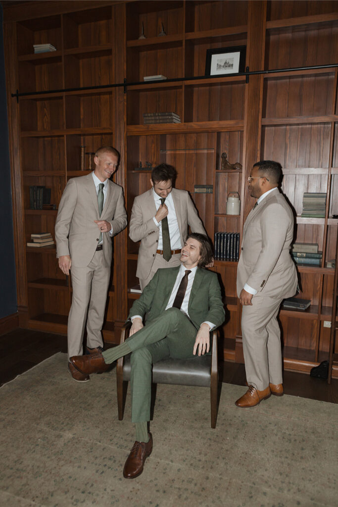 Four men in suits are in a room with wooden bookshelves. Three are standing; one in a green suit is sitting in a chair, smiling.