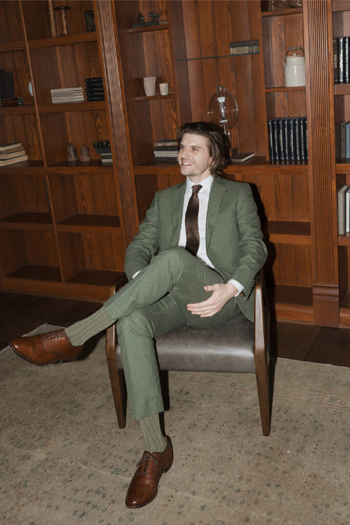 Man in a green suit sitting in a chair, smiling, surrounded by wooden bookshelves with books and decor items in The Grand Lady Austin.