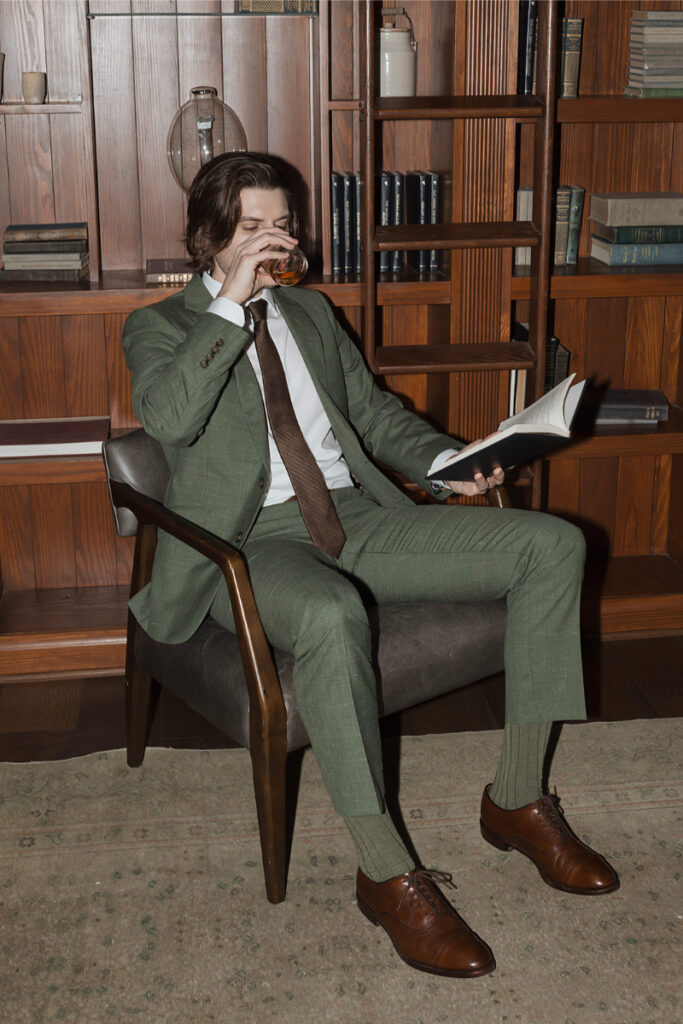 A person in a green suit sits in a chair drinking from a glass while reading a book, surrounded by wooden shelves with books in The Grand Lady Austin. 