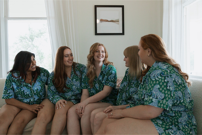 Five people sitting on a couch, wearing matching green patterned outfits, smiling and talking in a well-lit room.