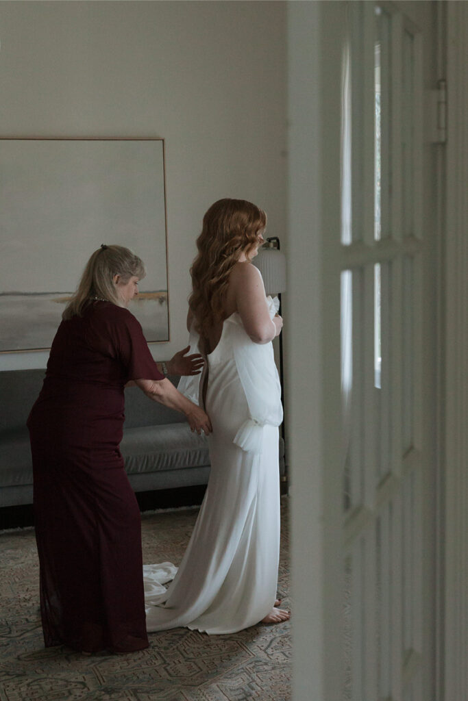 A woman in a burgundy dress helps another woman adjust her white dress in a room with a sofa and painting.