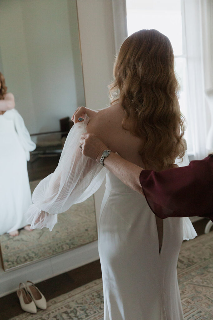 A woman with wavy hair adjusts her white dress in front of a mirror, assisted by someone in a maroon top. A pair of white shoes rests on the floor nearby.