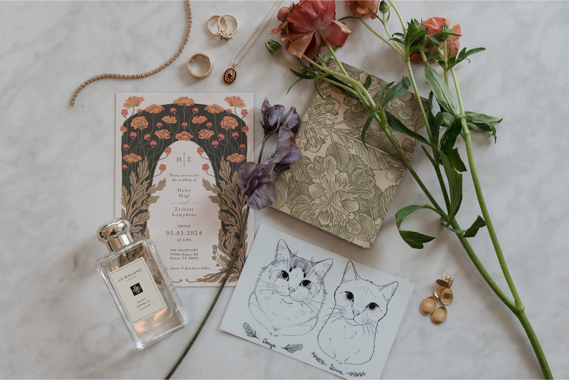 Wedding invitation set with floral designs, a perfume bottle, rings, a bracelet, flowers, and a card with cat illustrations on a marble surface.