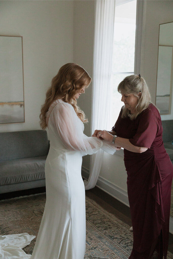 A woman in a white dress stands as another woman adjusts her sleeve in a room with a gray sofa and two paintings.