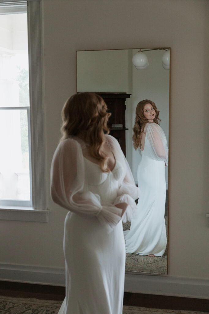 A woman in a white dress looks at her reflection in a mirror, smiling. She stands in a room with a window and simple decor.