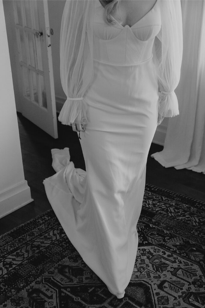 Person in a white bridal gown with pleated sleeves stands on a patterned rug near a door.