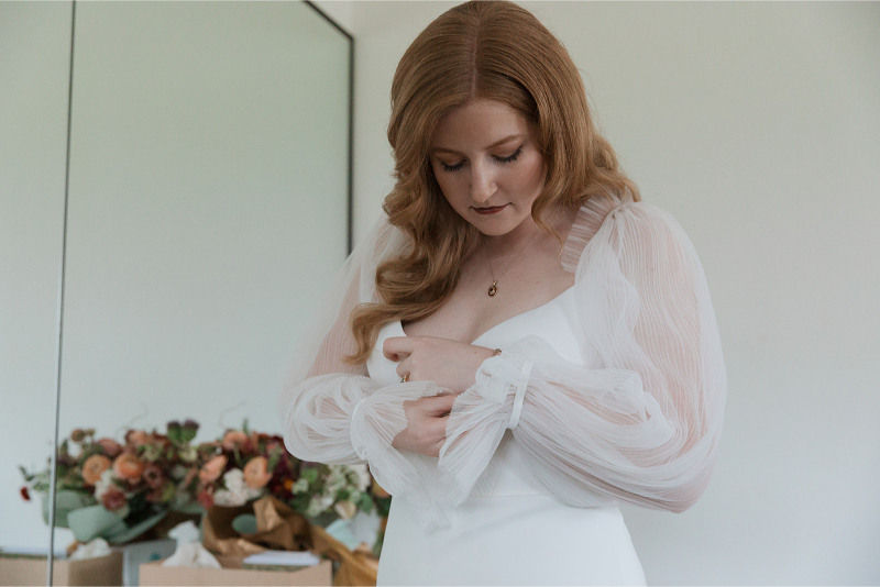 A woman in a white dress adjusts her sleeve. A bouquet of flowers is visible in the background.