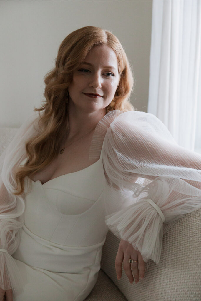 Woman with long red hair wearing a white dress sits on a couch, looking to the side with a slight smile.